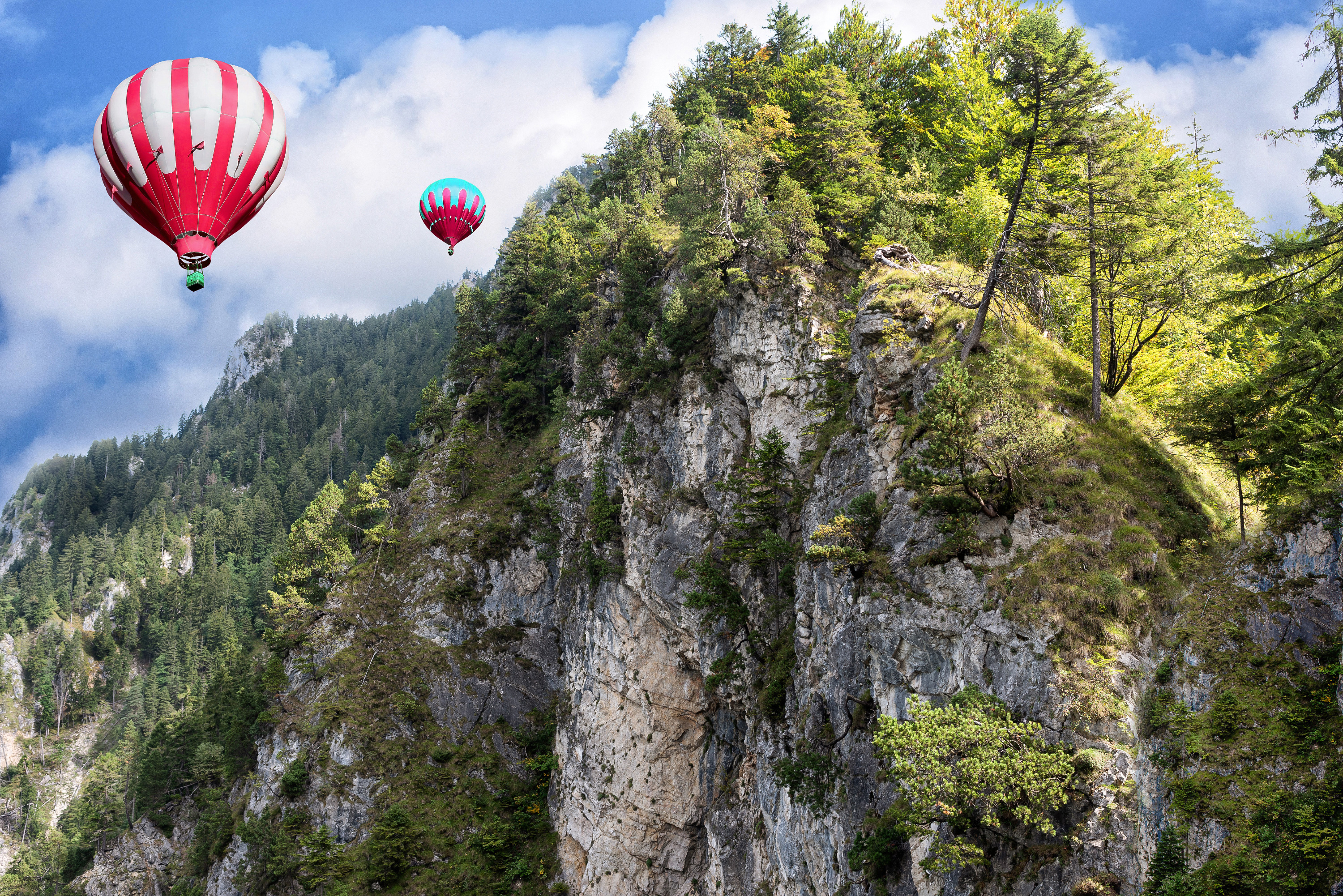 Red and white hot air balloons on background of high Alps mountains wth green forest under blue cloudy sky.